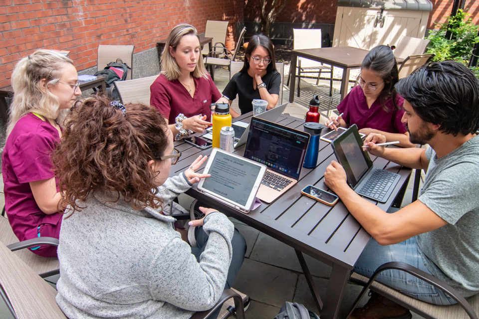 Six students outside at a table with laptops open, discussing coursework.