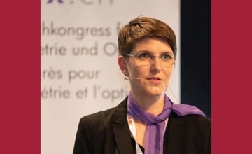 Nina Mueller has short hair and glasses, wearing a purple neck scarf, is speaking at a conference.