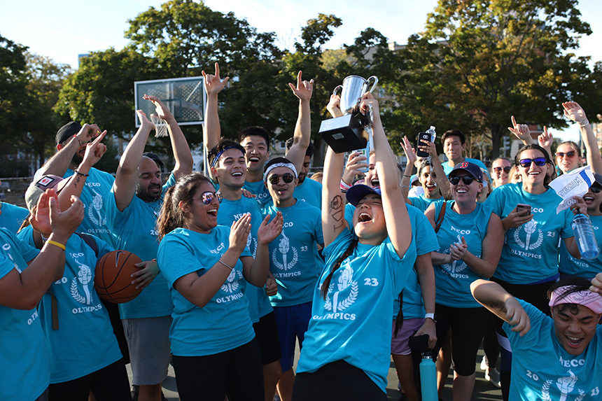 A group of students wearing blue t-shirts celebrate a win at student Olympics by holding up championship trophy and cheering