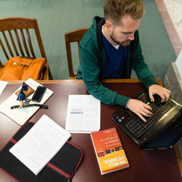 Overhead shot of male student wearing green sweatshirt and blue scrubs at laptop in library