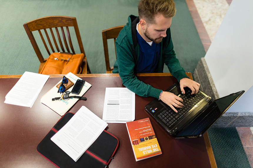 Overhead shot of male student wearing green sweatshirt and blue scrubs at laptop in library