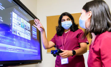 Two students wearing scrubs and masks in front of large computer monitor