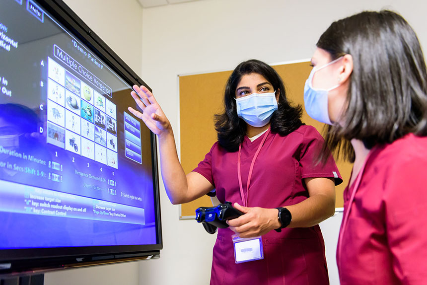 Two students wearing scrubs and masks in front of large computer monitor