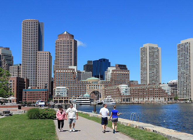 People walking along the harbor with tall buildings of Boston skyline in the background.