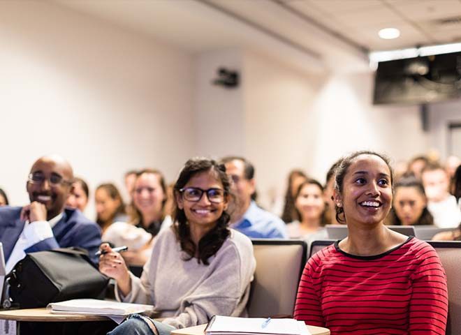 Two students in a lecture hall with many students behind them in the background.