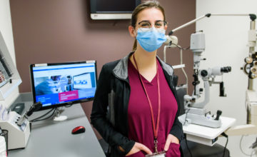 person smiling at camera with hands in pocket and standing in front of eye exam equipment