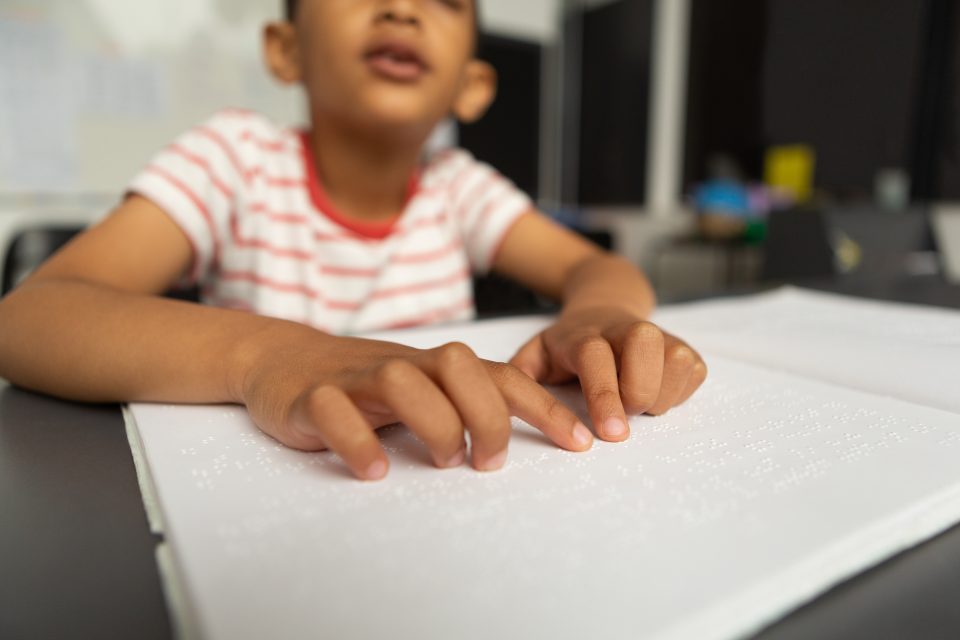 young child wearing a white t-shirt with red stripes reads pages printed in braille with his hands